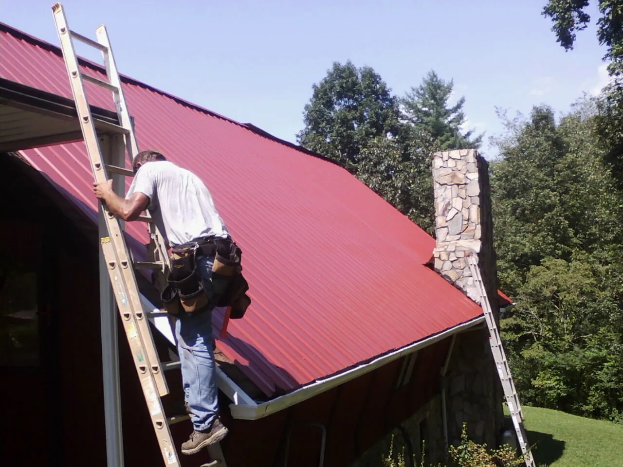 Roofer on a ladder beside a red metal roof and stone chimney — Soffit & Fascia Repair by Roofing Brothers in Albion